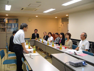 座っている8名の人物と立って発言している三浦部長